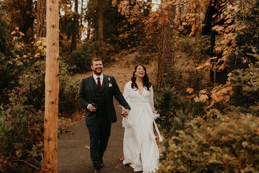 A newlywed couple smiles as they hold hands and walk down the wooded path at their venue. Image by Maya Lora Photography.