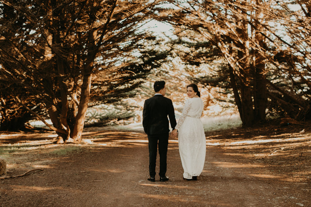 An image by Maya Lora Photography of a bride and groom holding hands and walking down a path with the bride looking back over her shoulder.
