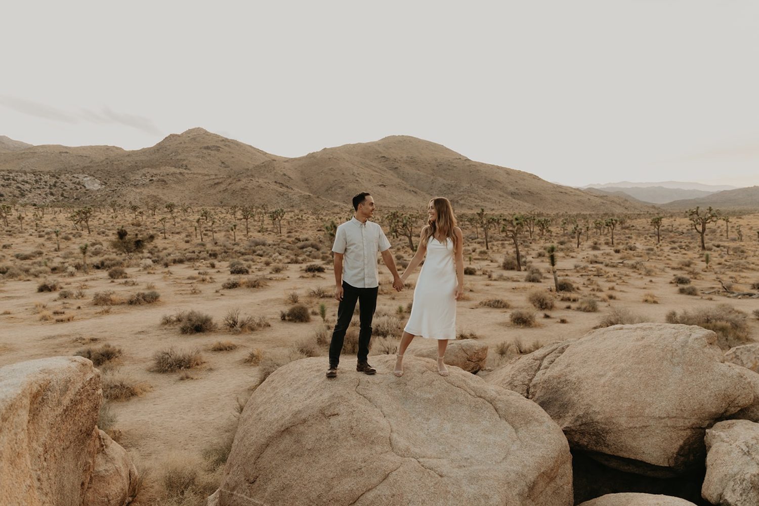 A couple hold hands while standing on a rock during their Joshua Tree engagement shoot.