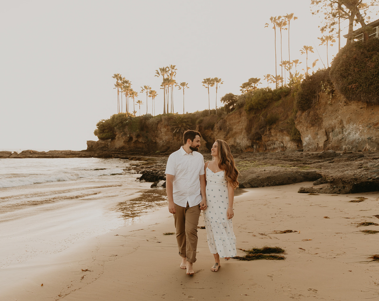 A couple holds hands while walking along Laguna Beach during their surprise proposal by Maya Lora Photo