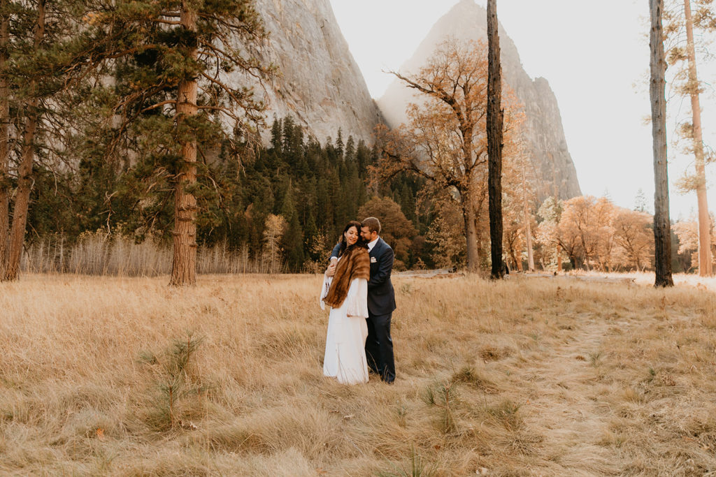Groom embraces bride from behind as they stand in a field of Yosemite National Park