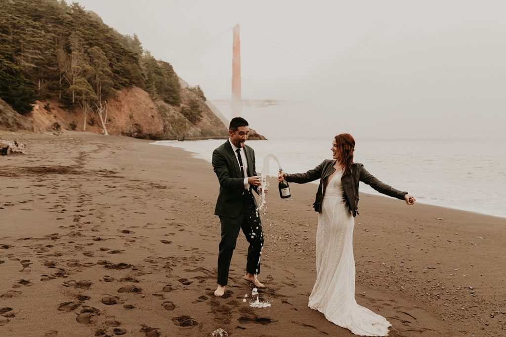 Bride and groom celebrate their nuptials by opening a bottle of champagne on a San Francisco beach; photo by Maya Lora Photography