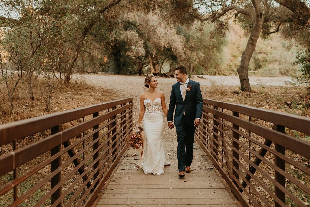 Bride and groom smile while holding hands and walking across a bridge together after their San Diego wedding