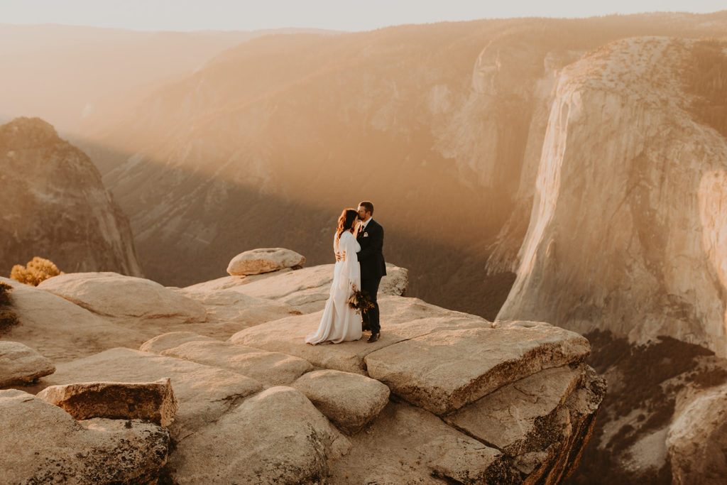 Bride and groom stand on a cliff overlooking Yosemite national park during their wedding photos by Maya Lora Photography
