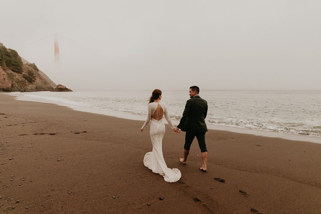 Bride and groom hold hands while walking along the beach during their San Francisco destination wedding